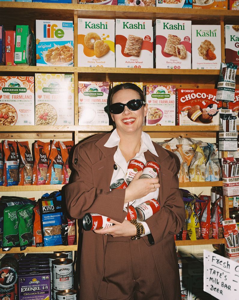 Woman in a brown suit hugging Campbell's cans inside a bodega