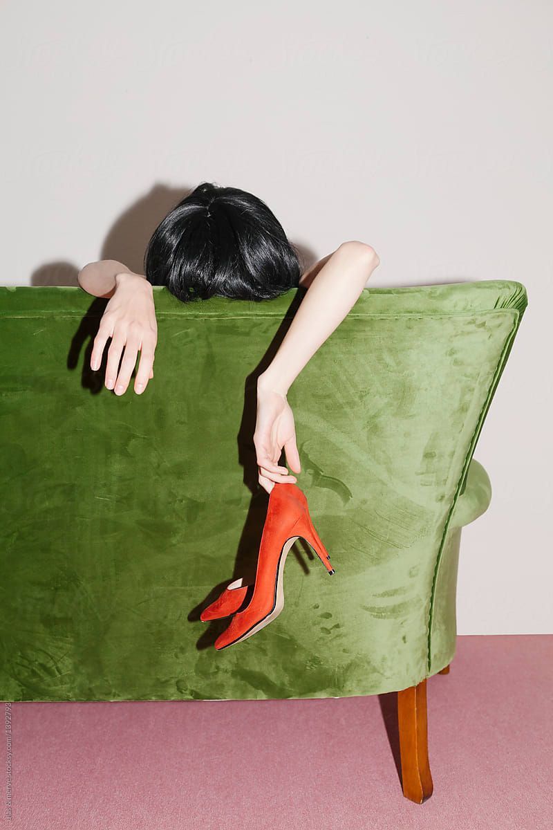 Headless figure draped over a green velvet sofa holding a red heel